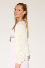 Woman posing confidently, wearing a white knit cardigan over a pink satin dress. She has long hair, hoop earrings, and a pearl necklace with a heart charm.