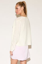 Woman posing confidently, wearing a white knit cardigan over a pink satin dress. She has long hair, hoop earrings, and a pearl necklace with a heart charm.