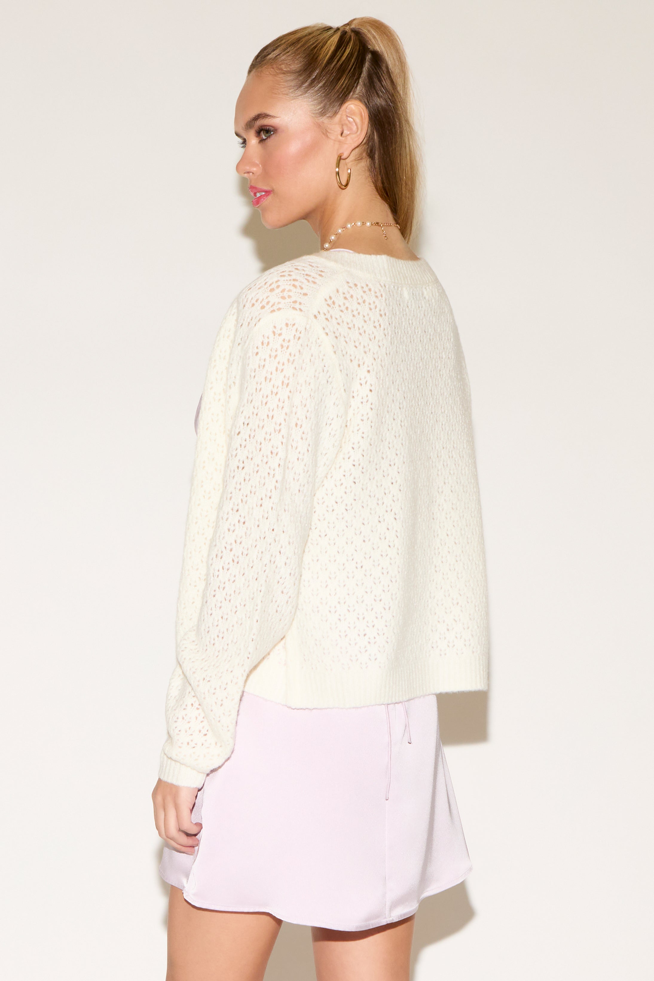 Woman posing confidently, wearing a white knit cardigan over a pink satin dress. She has long hair, hoop earrings, and a pearl necklace with a heart charm.