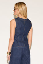A woman in a stylish, sleeveless denim jumpsuit and black sunglasses poses confidently. She holds a black handbag and wears dark heels, exuding modern elegance.
