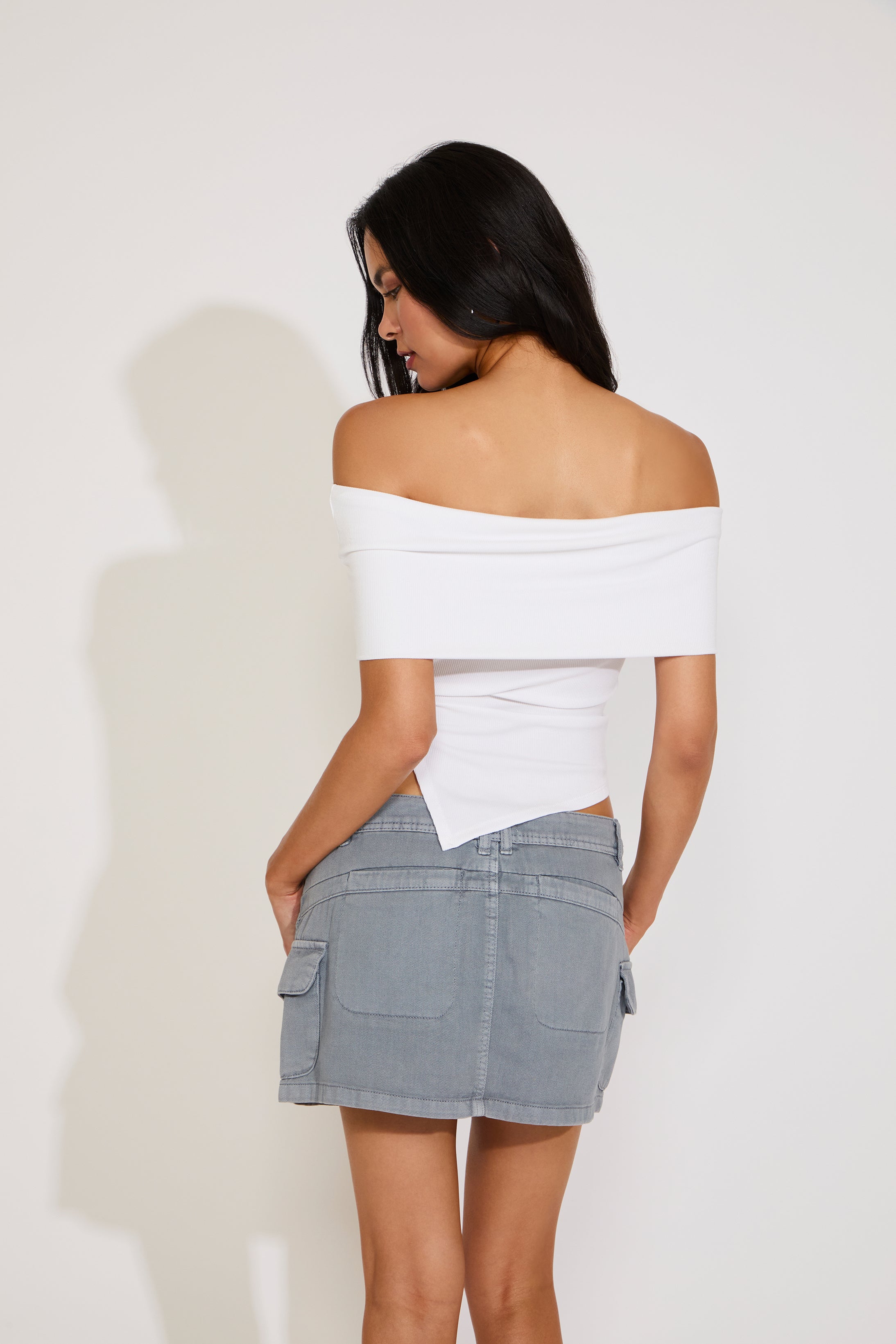 Woman wearing a white off-shoulder top and gray skirt against a plain background