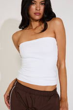 Woman wearing a white strapless top and brown pants against a plain background
