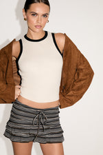 Woman wearing a white tank top with black trim, brown jacket, and striped shorts on a white background