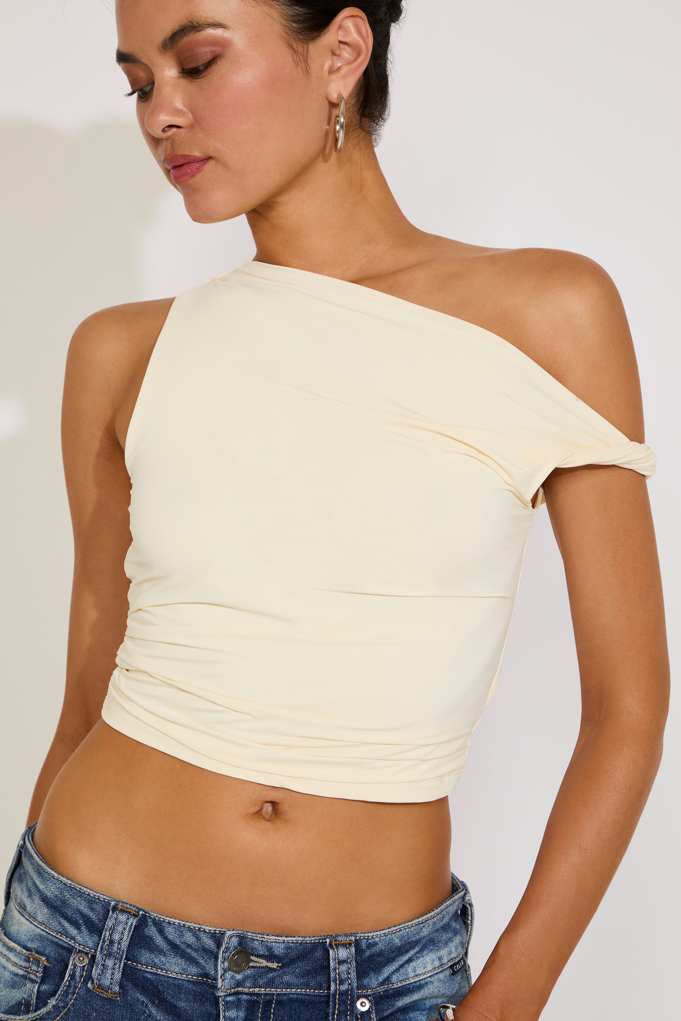 Woman wearing a cream-colored one-shoulder top and blue jeans on a plain background