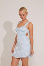 A woman standing wearing a sleeveless blue floral lace mini dress with a sweet neckline and lace trim.