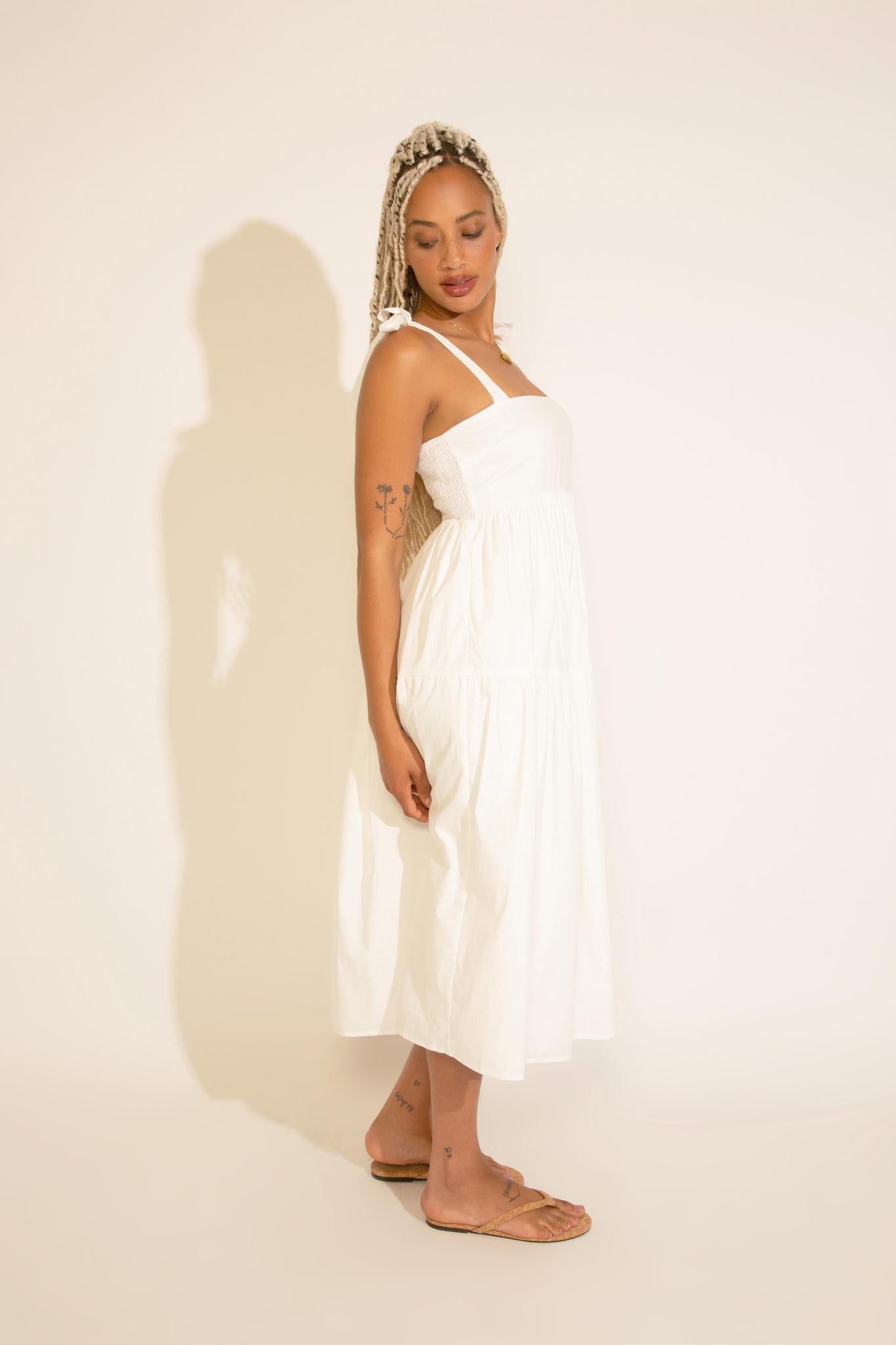 A woman standing wearing a white square neck midi dress with tied straps, a smocked bodice, and an A-line silhouette.