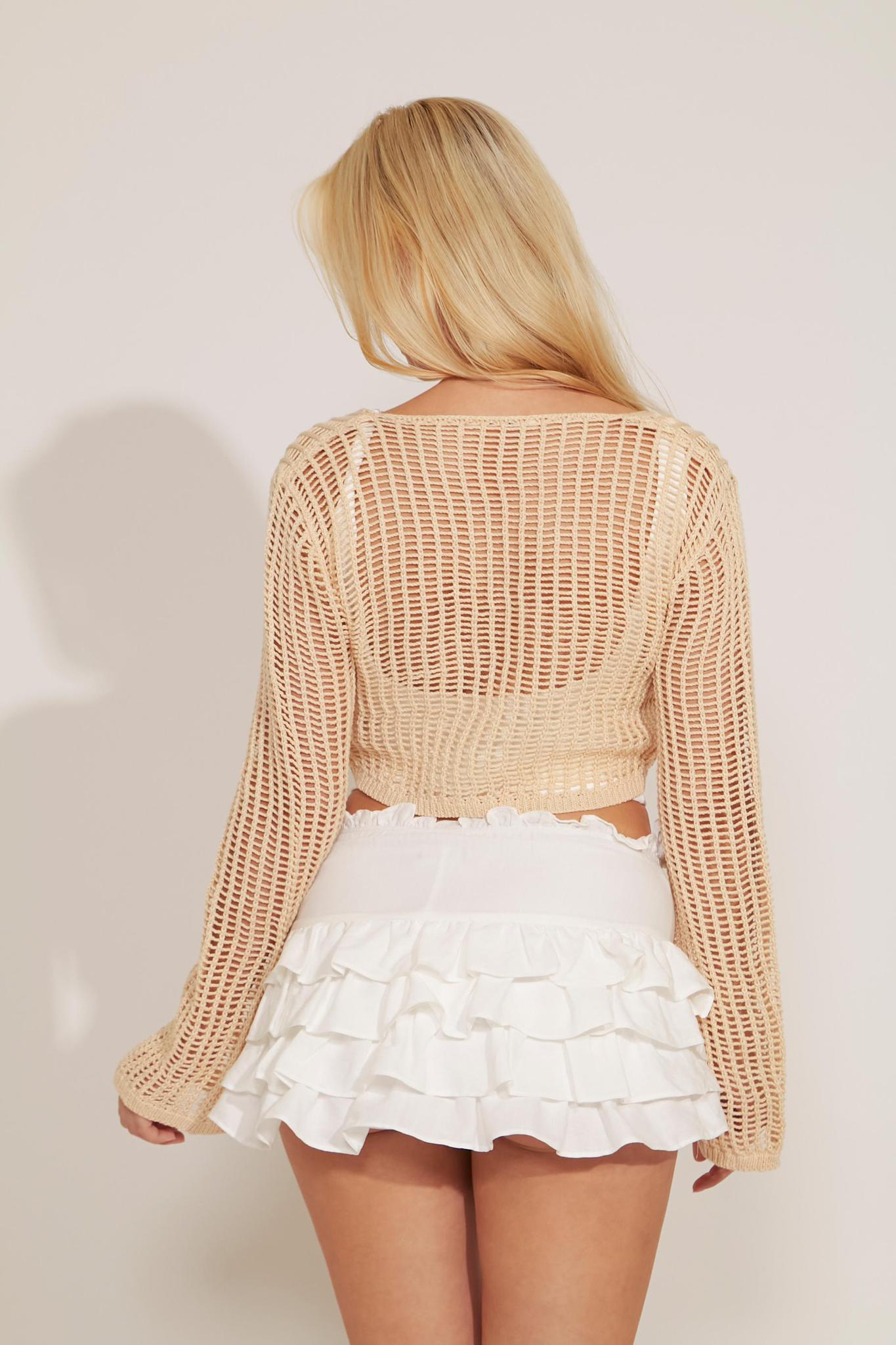 A woman wearing a beige crochet front tie-up cropped top with long sleeves, paired with a white mini skirt.