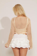 A woman wearing a beige crochet front tie-up cropped top with long sleeves, paired with a white mini skirt.