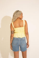 A woman wearing a sleeveless yellow peplum top with ruched detailing and an elastic banding, paired with blue denim shorts.