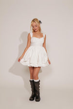 A woman is wearing a white empire waist ruffled mini dress with tie details and black boots.