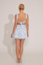 A woman standing wearing a sleeveless blue floral lace mini dress with a sweet neckline and lace trim.