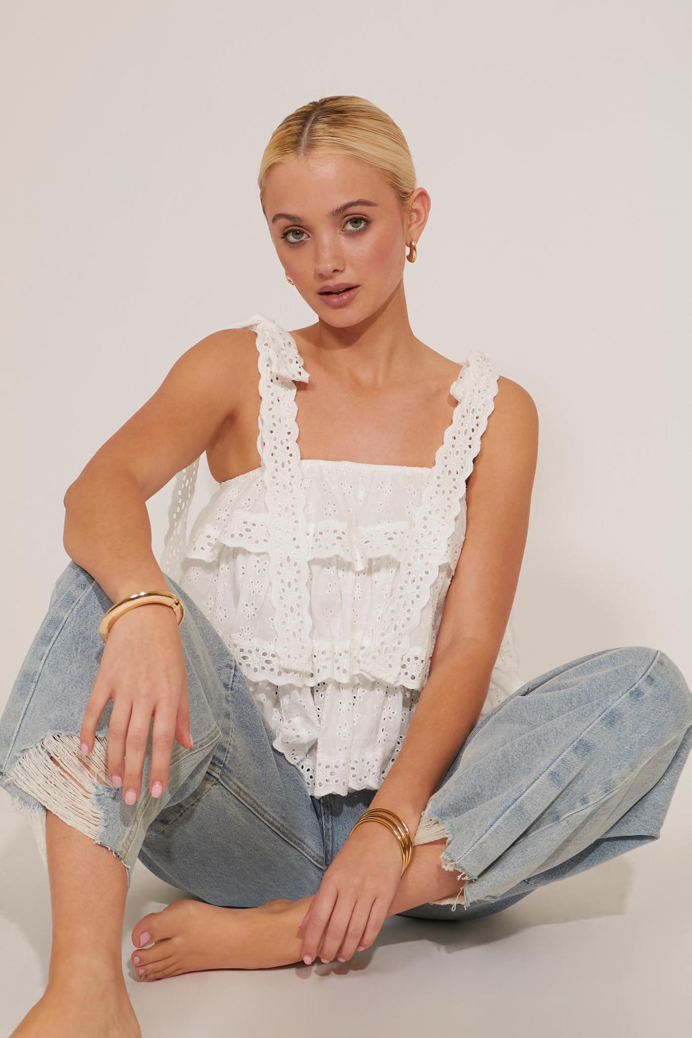 A woman sitting on the floor wearing a white tiered tank top with lace straps and floral embroidery, paired with blue denim shorts.