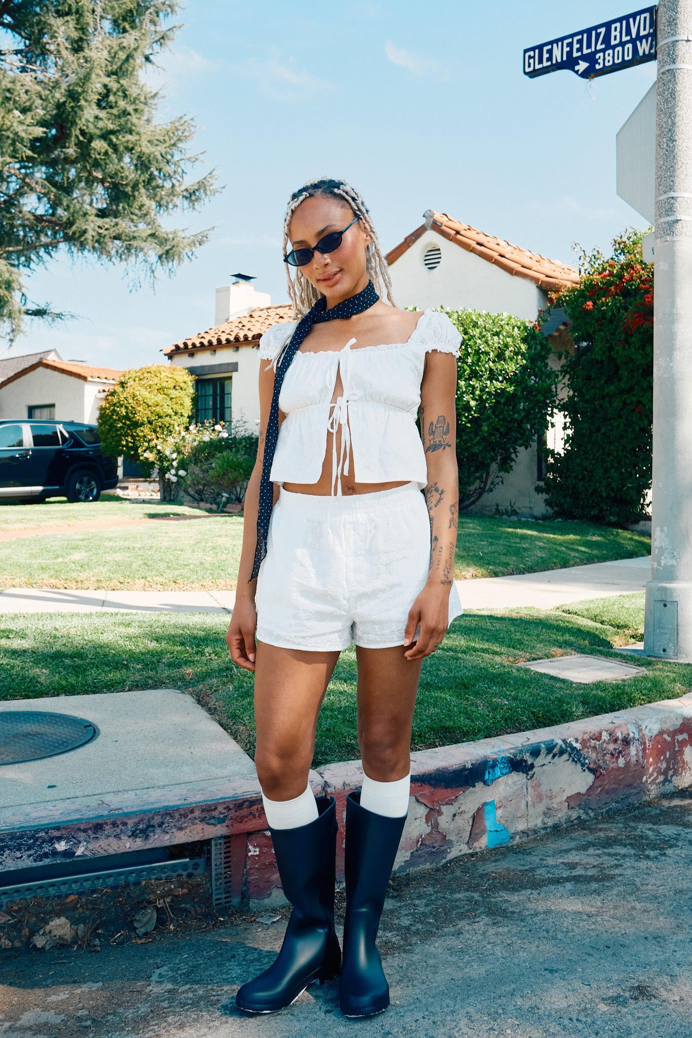 A woman wearing a white cap sleeve square neck open top with shorts, sunglasses, and a neck piece.