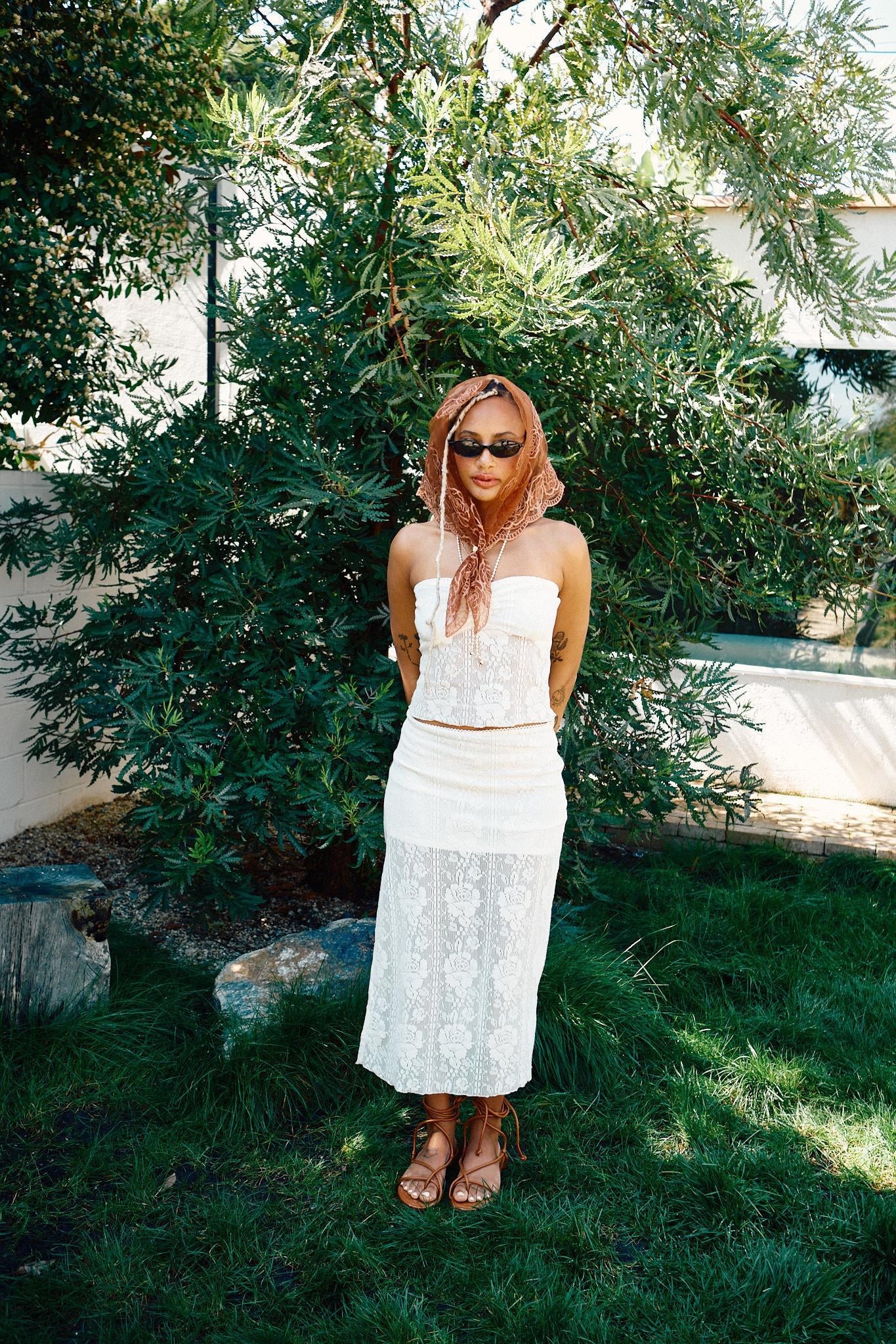 A person wearing a white maxi skirt with a rose pattern and lace detailing, standing on grass with trees and shrubs in the background.