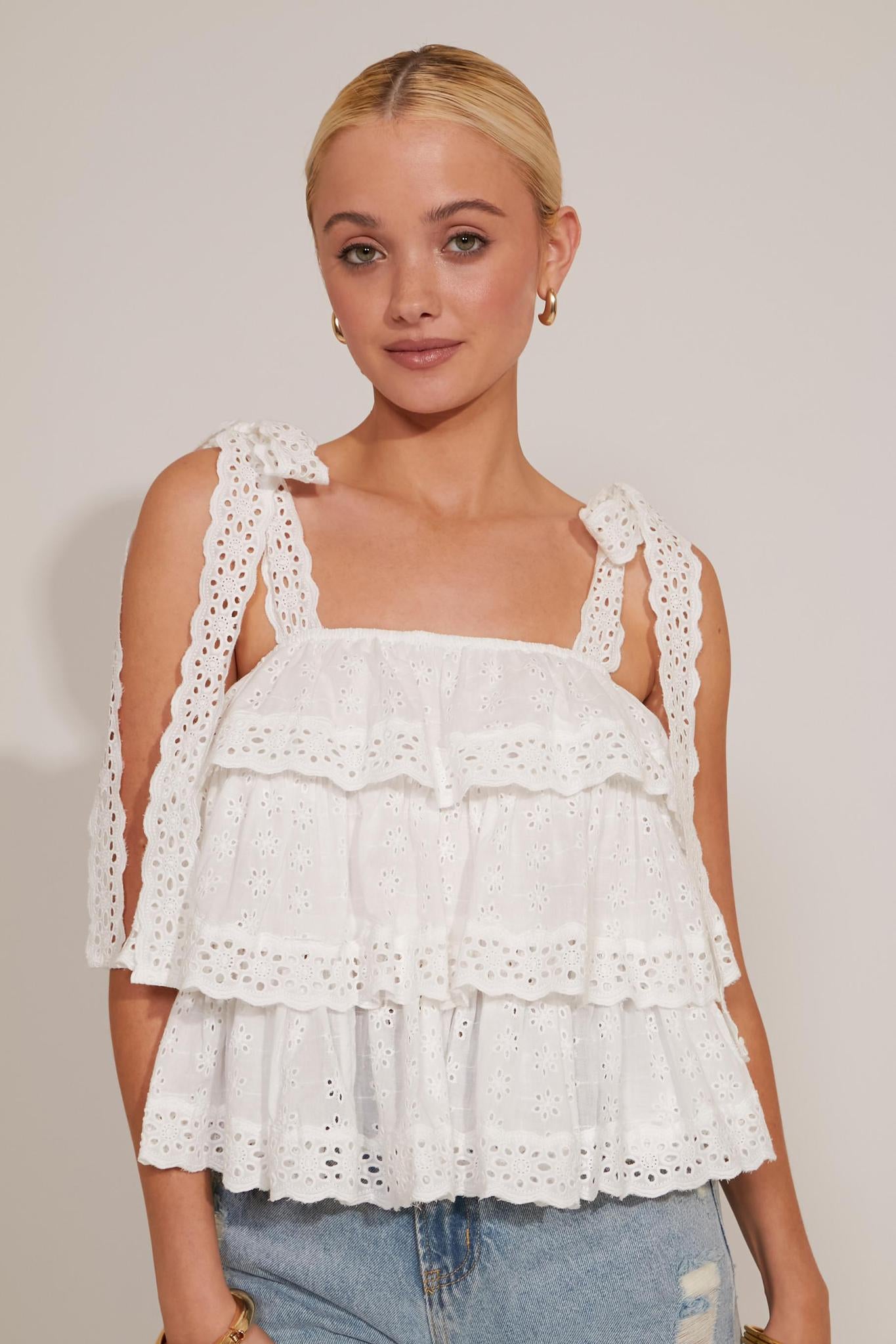 A woman wearing a white tiered top with embroidered details and lace straps, paired with blue ripped jeans.