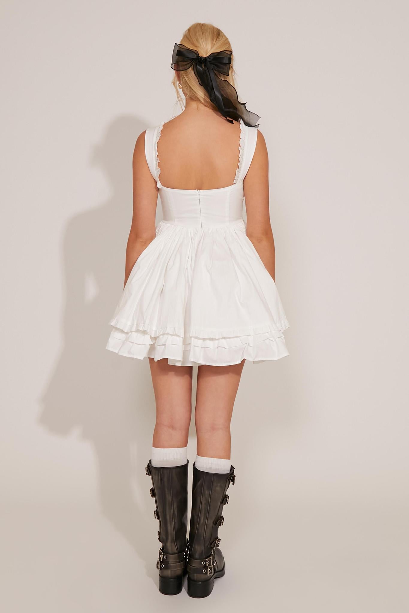 A woman is wearing a white empire waist ruffled mini dress with tie details and black boots.