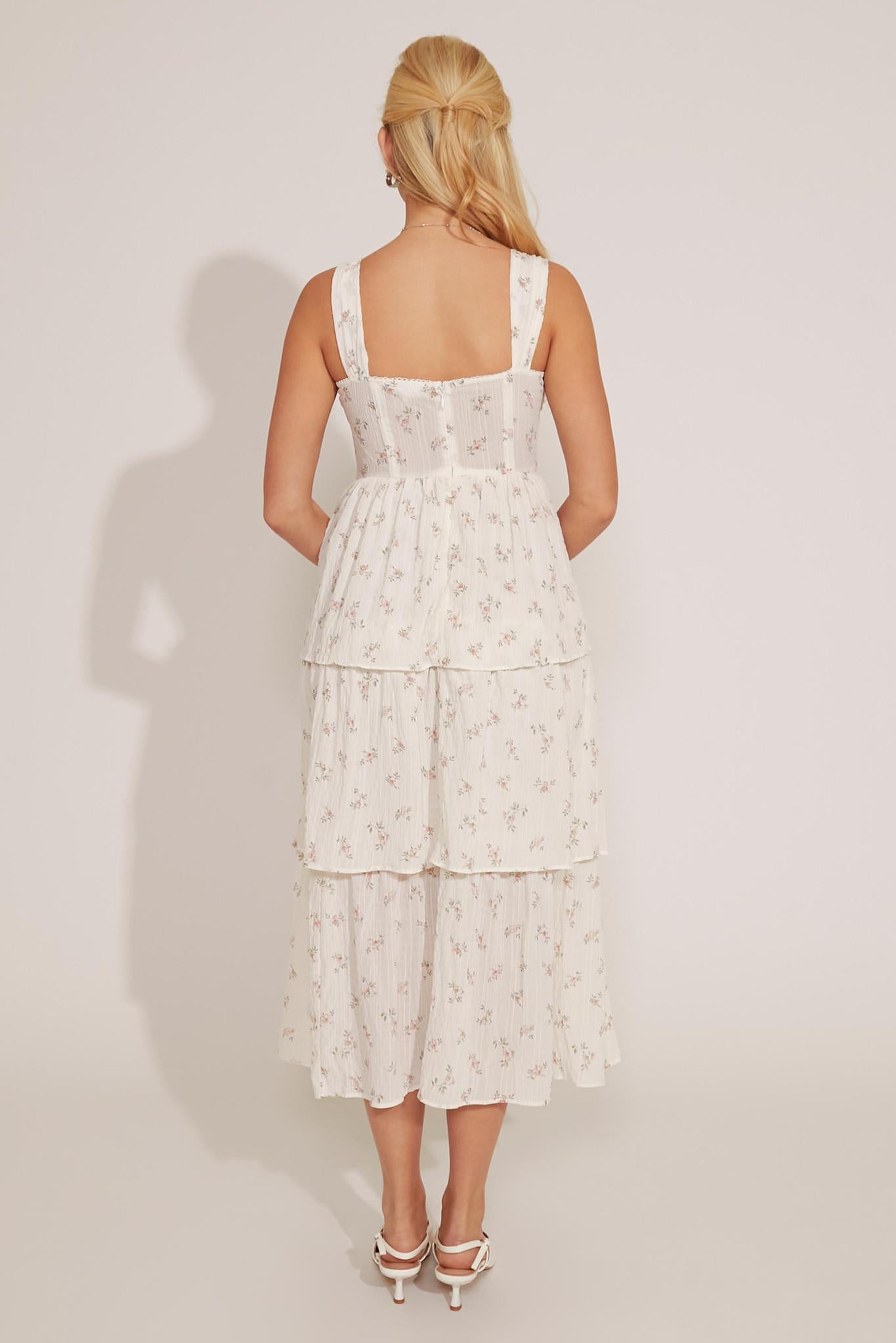 A woman modeling a floral print midi dress with layered ruffles and bone wire structure, wearing white shoes.